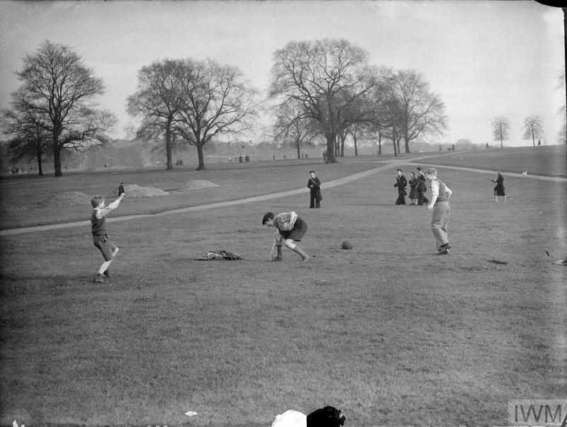 Boys, including a Boy Scout, enjoy a game of football in the sunshine in Hyde Park. A naval cadet and several other children can be seen in the background, watching from a distance.